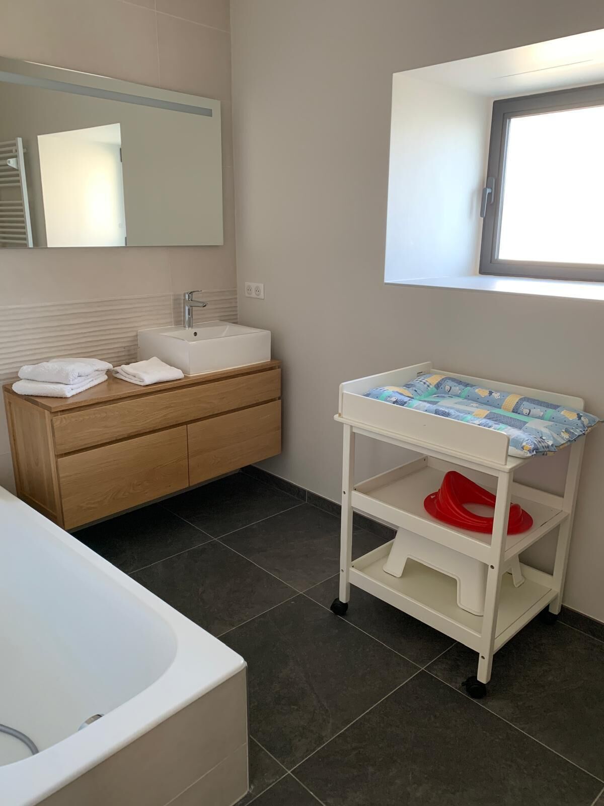 Une salle de bain avec un lavabo blanc, un meuble-lavabo en bois, une étagère blanche sur laquelle est posé un pot de chambre rouge, et un grand miroir.