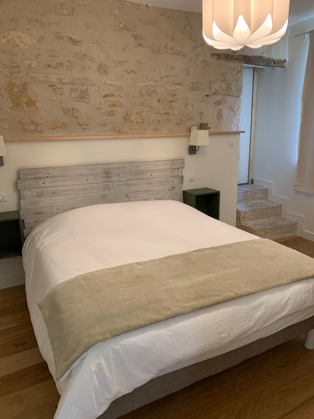 Un dormitorio con cabecera de madera, una cama grande con ropa de cama blanca y un edredón beige, y una lámpara de pared.