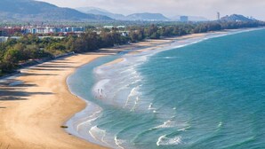 A coastal beach with a sandy shoreline, a clear blue sea, and a backdrop of hills and buildings. - 75/2 Residence Hua Hin (Hua Hin)