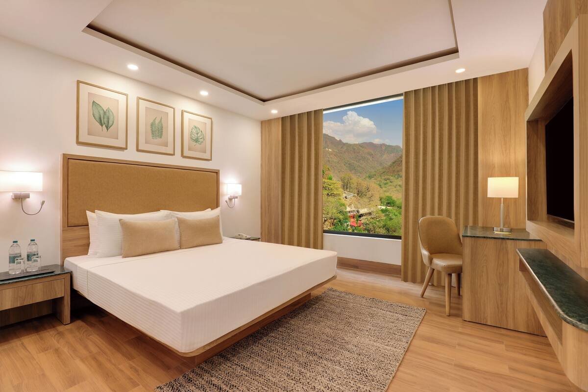 A modern hotel room with a large bed, a desk, and a view of a mountainous landscape.