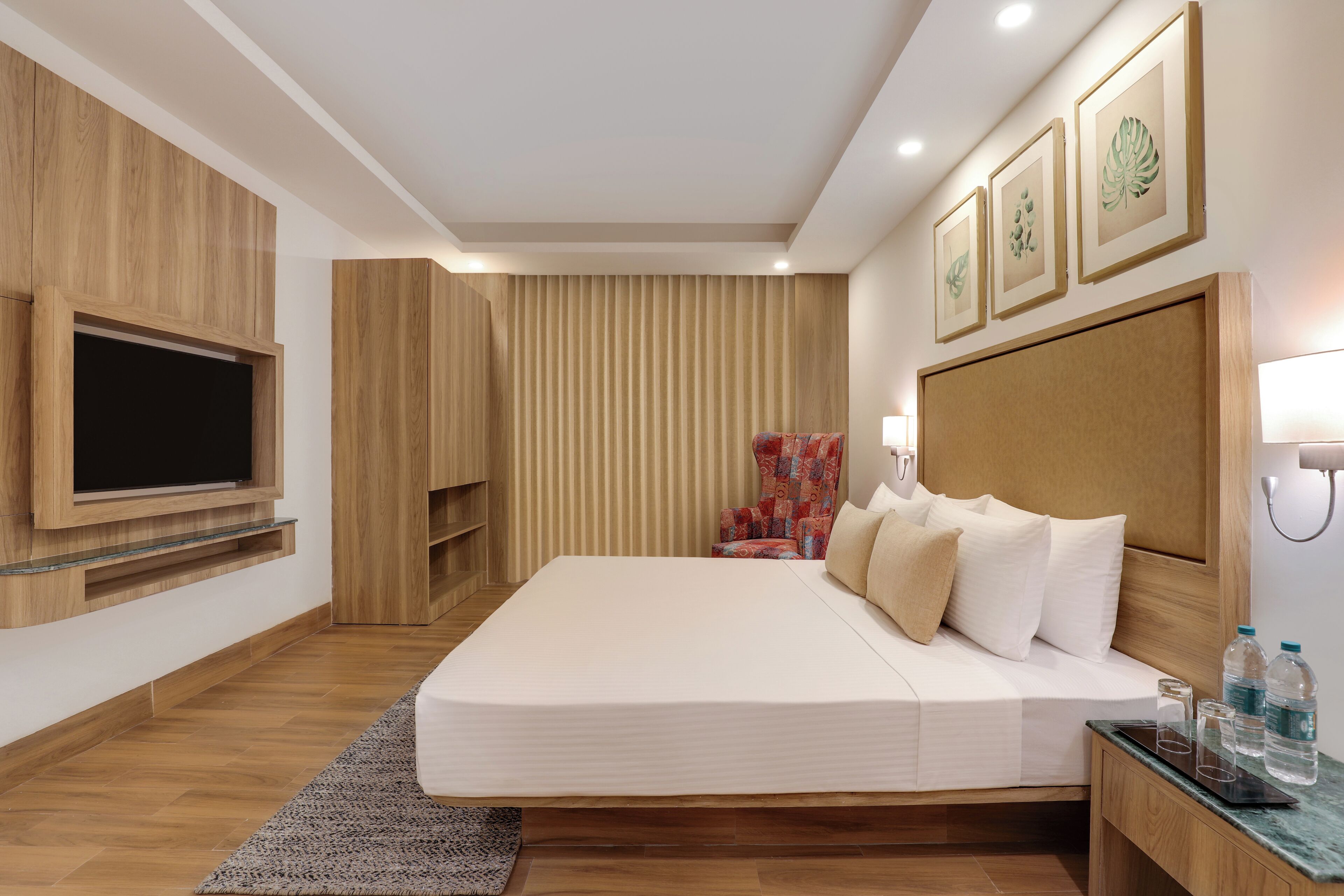 A modern hotel room with a large bed, a flat-screen TV mounted on the wall, a patterned armchair, and a wooden headboard.