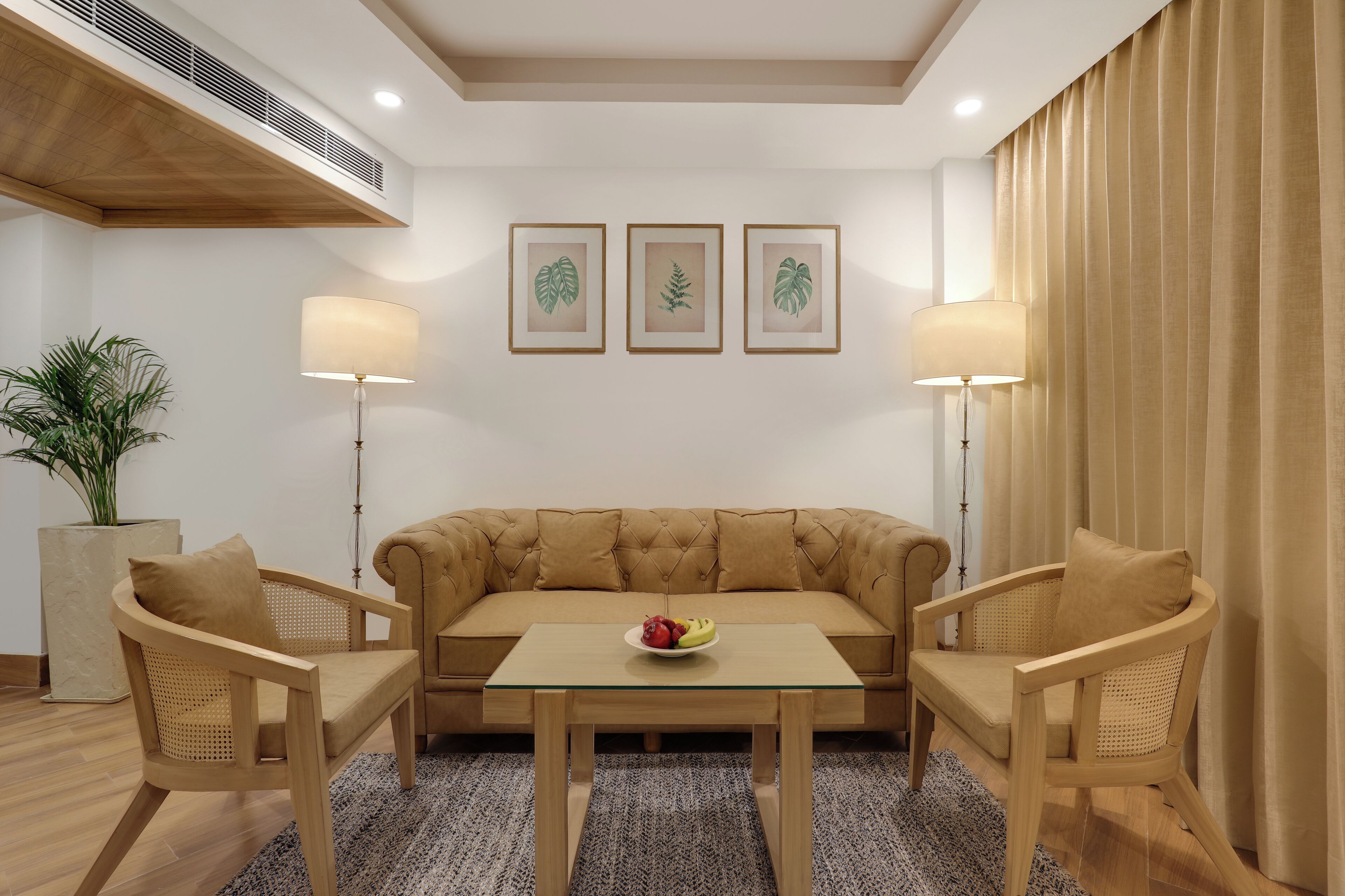 Een moderne woonkamer met een beige bank, twee rieten fauteuils, een glazen salontafel met fruit, en drie ingelijste botanische prints aan de muur.