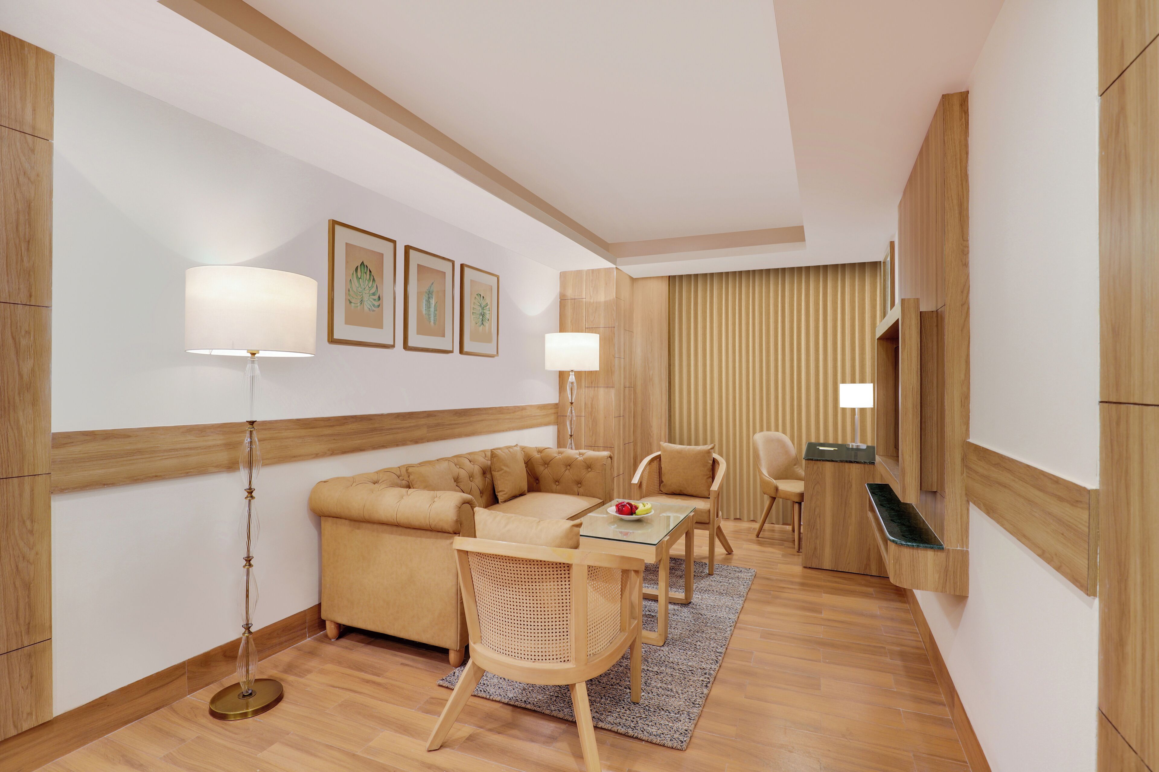 A modern hotel room with a sectional sofa, a glass-top dining table, two chairs, a floor lamp, and a small side table with a fruit bowl.