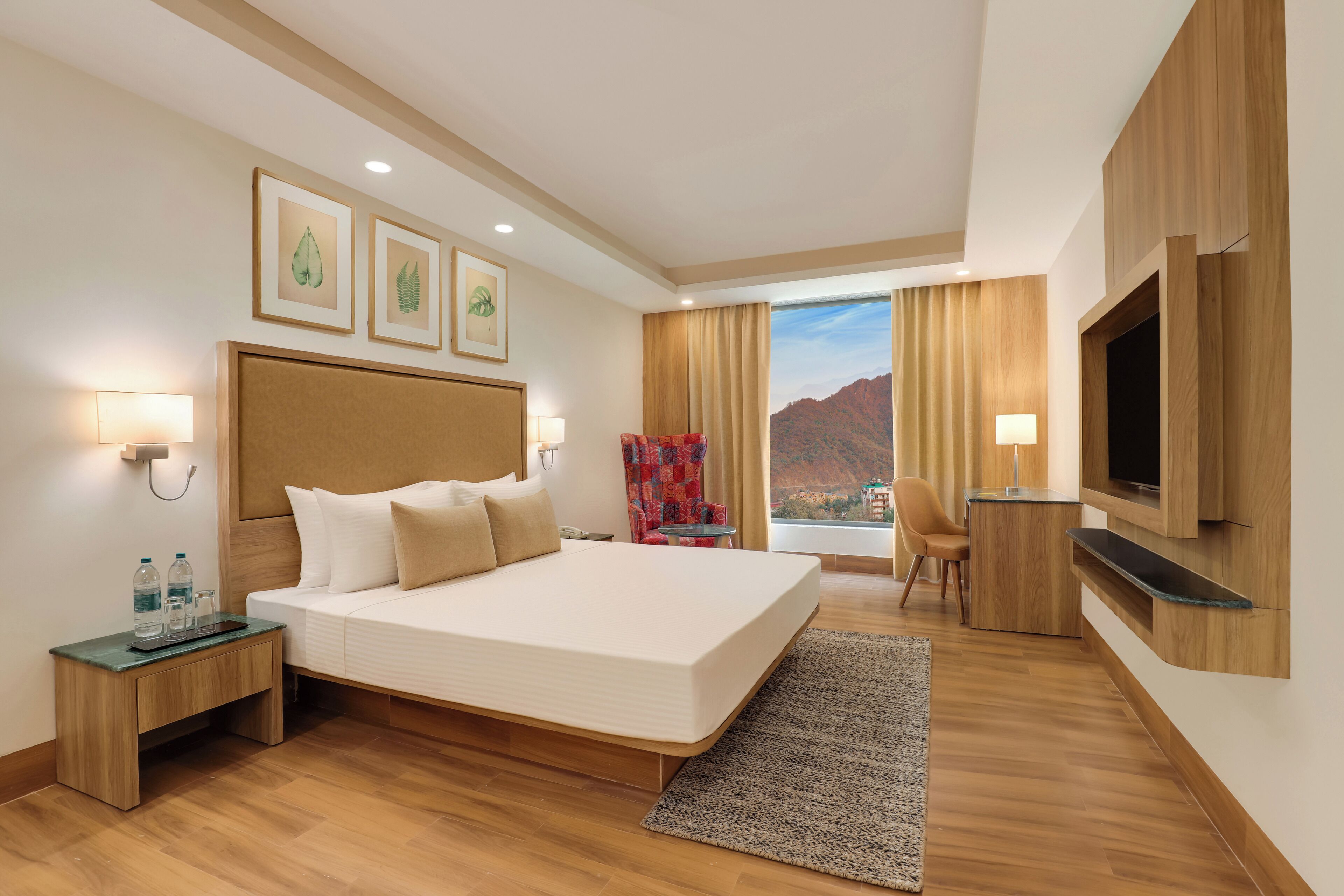 A hotel room with a large bed, a TV mounted on the wall, a desk with a chair, and a view of mountains through the window.