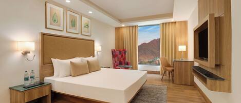A hotel room with a large bed, a TV mounted on the wall, a desk with a chair, and a view of mountains through the window.