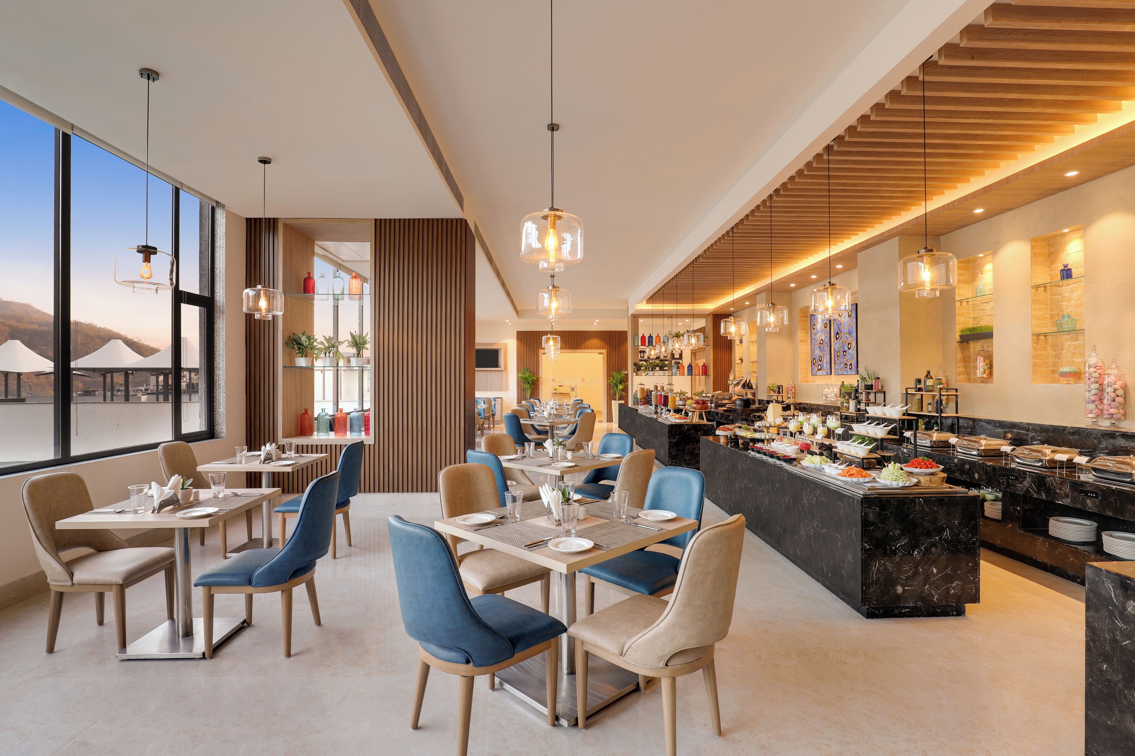 A modern dining area with tables and chairs, a buffet counter, and large windows offering a view of the outdoors.