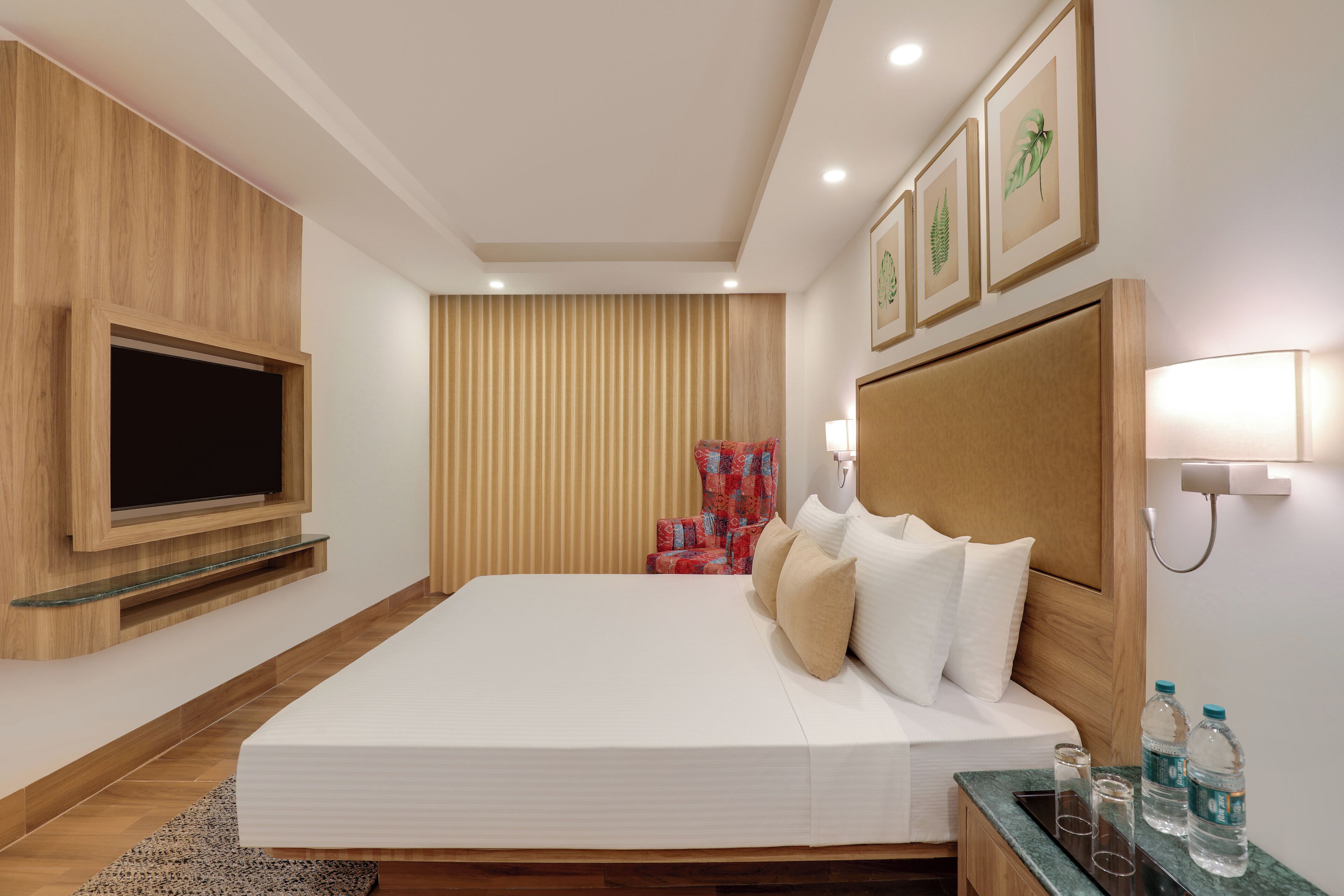 Ein modernes Hotelzimmer mit einem großen Bett, einem flachbildfernseher an der Wand, einem Holz-Kopfteil und einem Nachttisch mit Wasserflaschen.