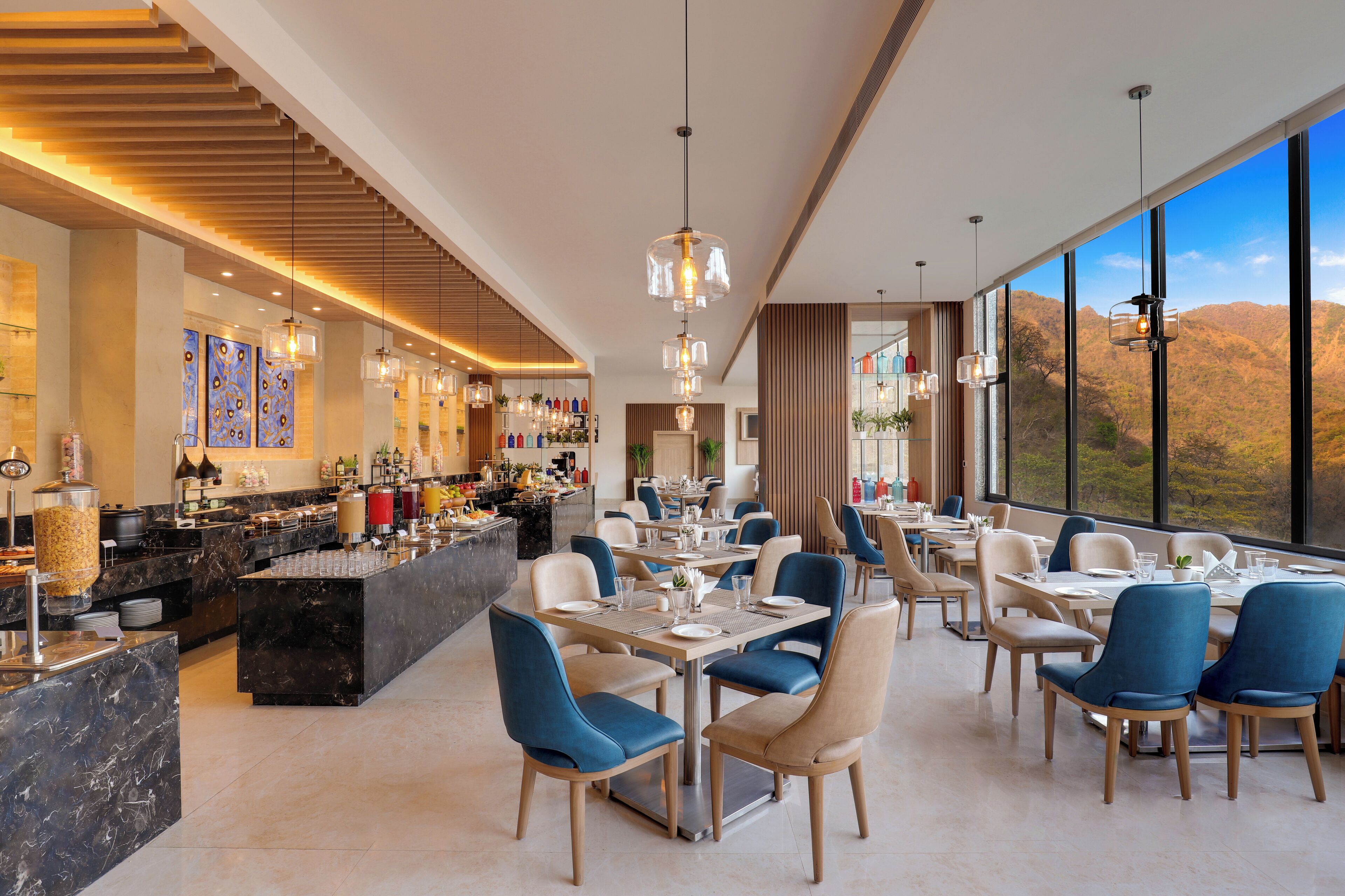 A modern restaurant with a long counter, multiple dining tables, and chairs. There is a bar area with various bottles and a large window offering a view of the hills.