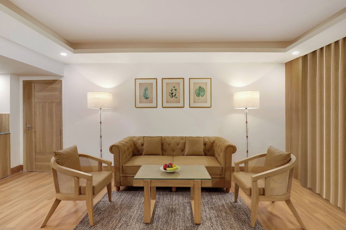 A modern living room with a beige sofa, two wooden chairs, a glass coffee table, and three framed botanical prints on the wall.