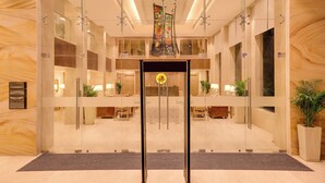 A modern hotel lobby with a reception desk, glass doors, and a central chandelier.