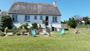 A two-story house with a sloped roof, a front porch, and a small garden with potted plants and outdoor furniture. - Location Maison Plein Pied 6 Personnes 800 ml de la Plage et du Port a Pied (Camaret-sur-Mer)