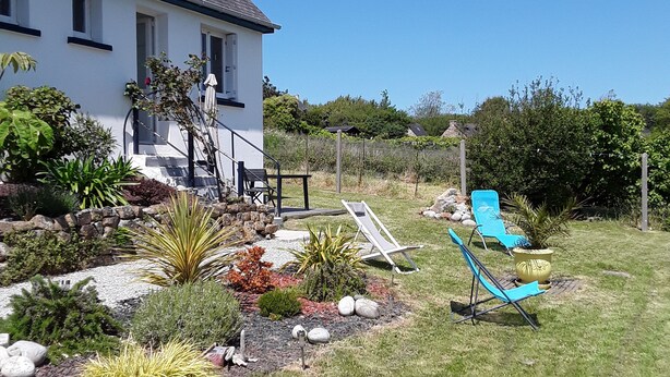 A house with a white exterior, a grey roof, and a small garden with various plants and a few outdoor chairs. - Location Maison Plein Pied 6 Personnes 800 ml de la Plage et du Port a Pied (Camaret-sur-Mer)
