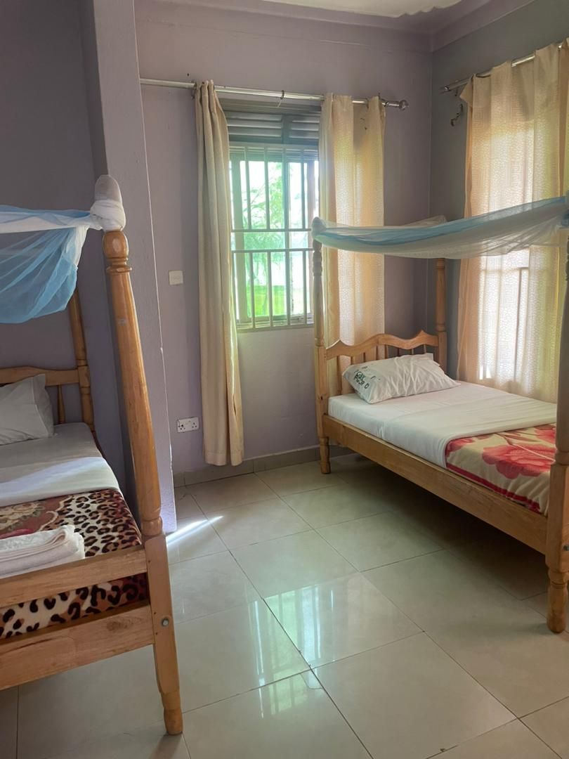 A room with two wooden beds, one with a canopy and one without, a window with curtains, and a tiled floor.