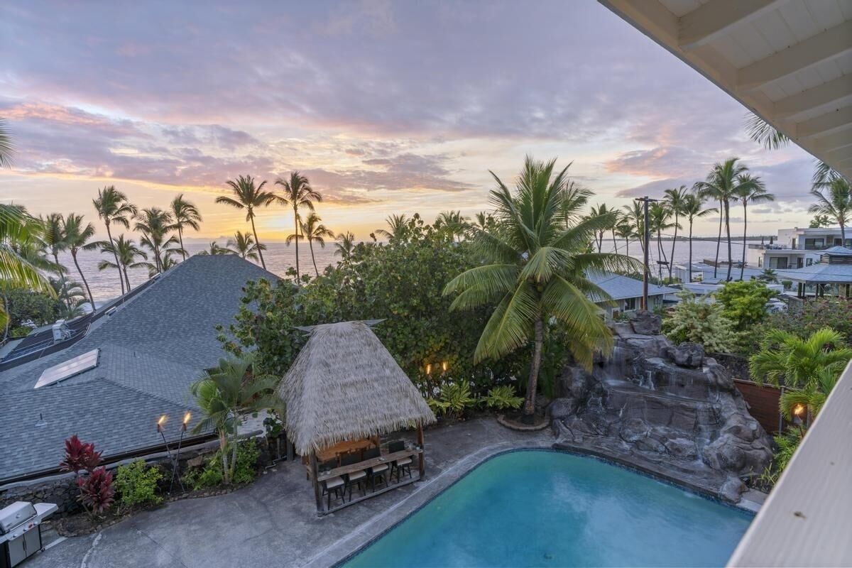 A pool area with a thatched-roof structure, palm trees, and a view of the ocean at sunset.