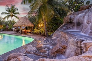 A pool with a waterfall, a thatched-roof structure, and lush tropical vegetation.