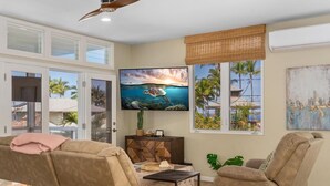 A living room with a large TV, beige sofas, a small table, and a window with a view of palm trees. - Big Island Aloha Pearl House 4 Bedroom Home (Kailua-Kona)