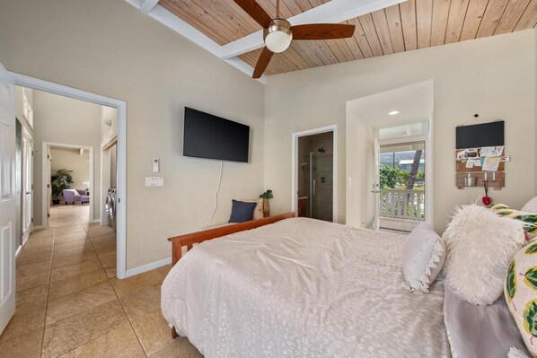 A bedroom with a large bed, a ceiling fan, a TV mounted on the wall, and a balcony door leading to an outdoor area.