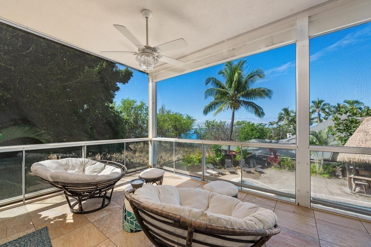 A spacious balcony with a glass railing, two cushioned chairs, a ceiling fan, and a view of palm trees and a thatched-roof structure.