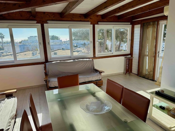 A room with a glass dining table, a covered sofa, and large windows offering views of buildings and trees.
