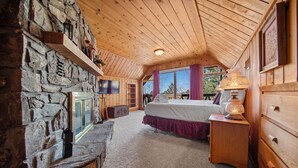 Une chambre confortable avec une cheminée en pierre, un plafond en bois et un lit orné d’une jupe rouge.
