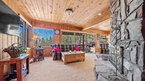 L’intérieur chaleureux d’un chalet avec des murs en bois, une cheminée en pierre, un canapé à carreaux et une grande fenêtre donnant sur la forêt.