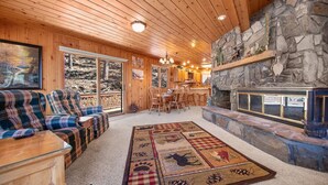L’intérieur chaleureux d’un chalet avec une cheminée en pierre, un canapé à carreaux et une grande fenêtre donnant sur une forêt.