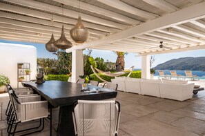 A spacious outdoor dining area with a large table, white wicker chairs, and hanging pendant lights.