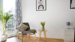 Un salon moderne avec un plancher en bois, un fauteuil gris, une petite table ronde ornée d’un vase de fleurs et une table de chevet blanche avec une lampe.