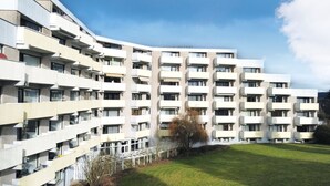 Un immeuble résidentiel moderne à plusieurs étages, doté de balcons, d’un toit plat et d’une cour verdoyante.