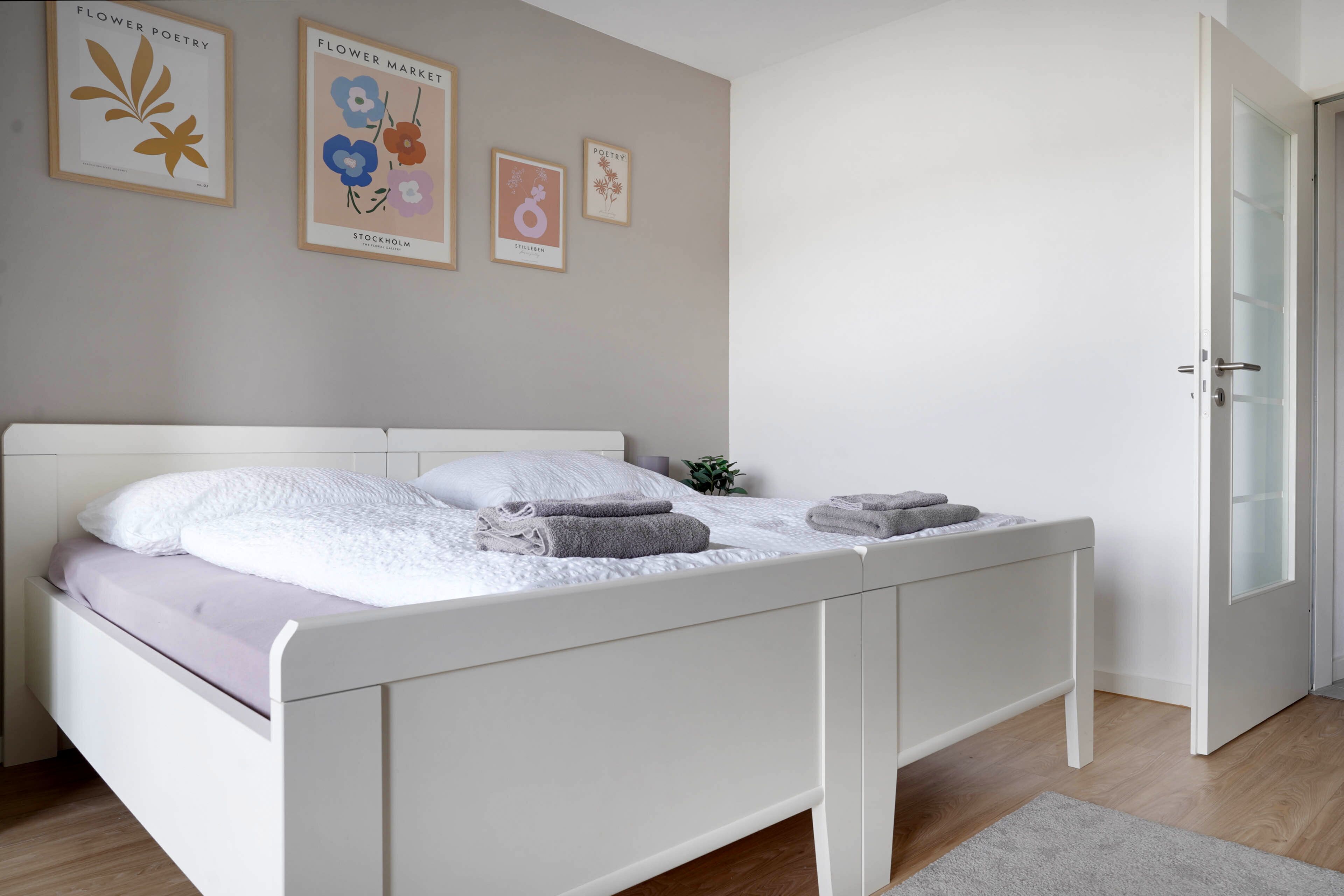 Ein ordentlich bezogenes Bett mit weißer Bettwäsche und zwei gefalteten Handtüchern, eine kleine Zimmerpflanze auf dem Nachttisch und Bilderrahmen an der Wand.