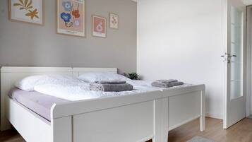 Ein ordentlich bezogenes Bett mit weißer Bettwäsche und zwei gefalteten Handtüchern, eine kleine Zimmerpflanze auf dem Nachttisch und Bilderrahmen an der Wand.