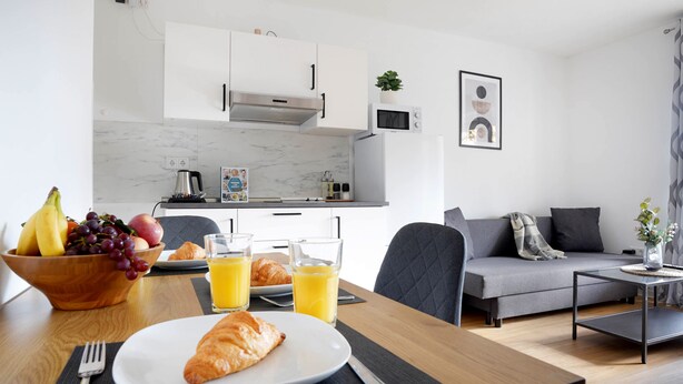 Une cuisine moderne avec des meubles blancs, un four à micro-ondes et une machine à café. On y trouve une table à manger avec un croissant, une assiette de fruits et des verres de jus d’orange. Un canapé gris et une petite table d’appoint avec un vase se trouvent dans l’espace salon.