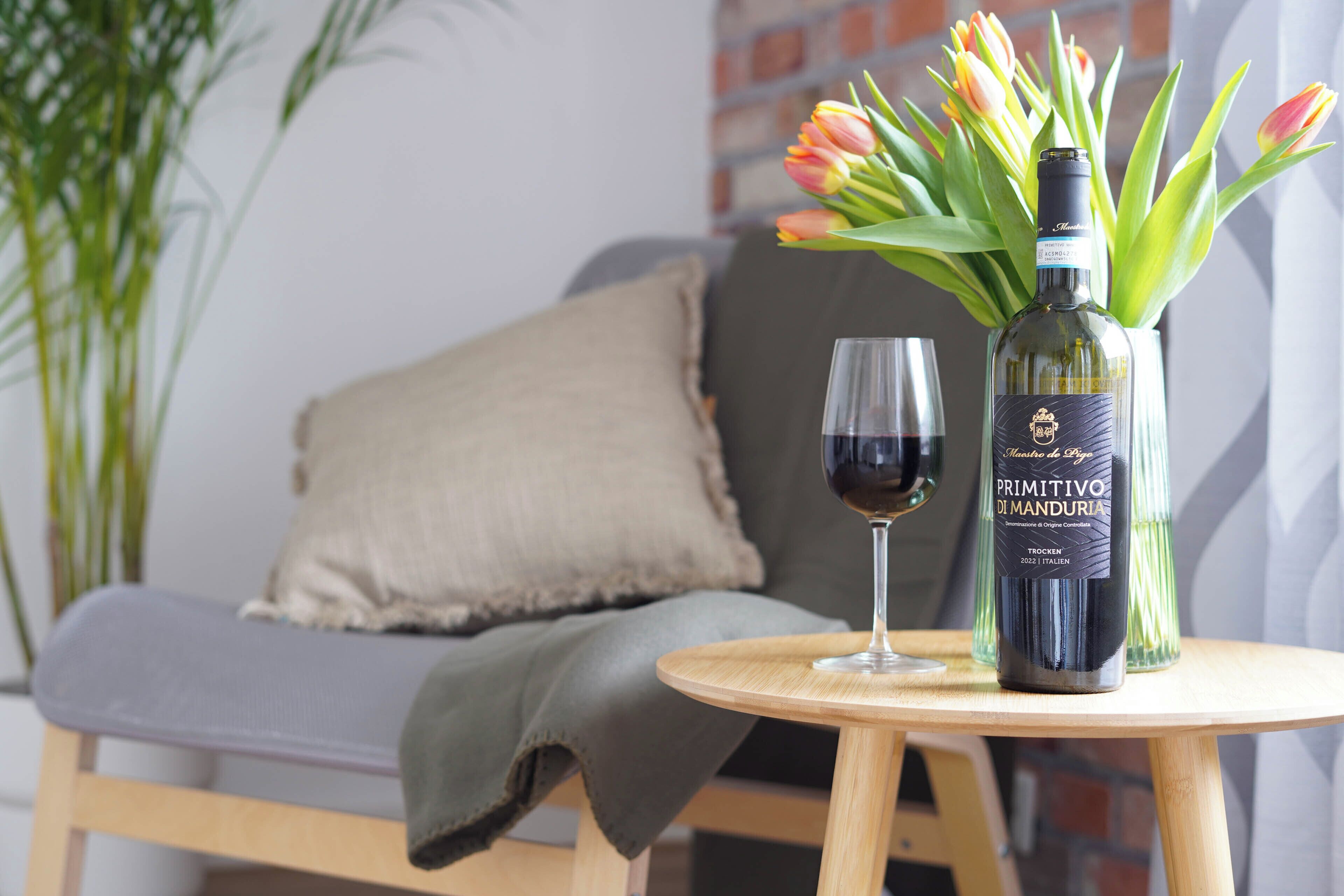 A bottle of wine and a glass on a small round table, with a cushion and a plant in the background.