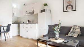 Une cuisine moderne avec des meubles blancs, un four à micro-ondes et un petit îlot central. Le salon est agrémenté d’une table basse en verre, d’une télécommande et d’un vase avec des fleurs.