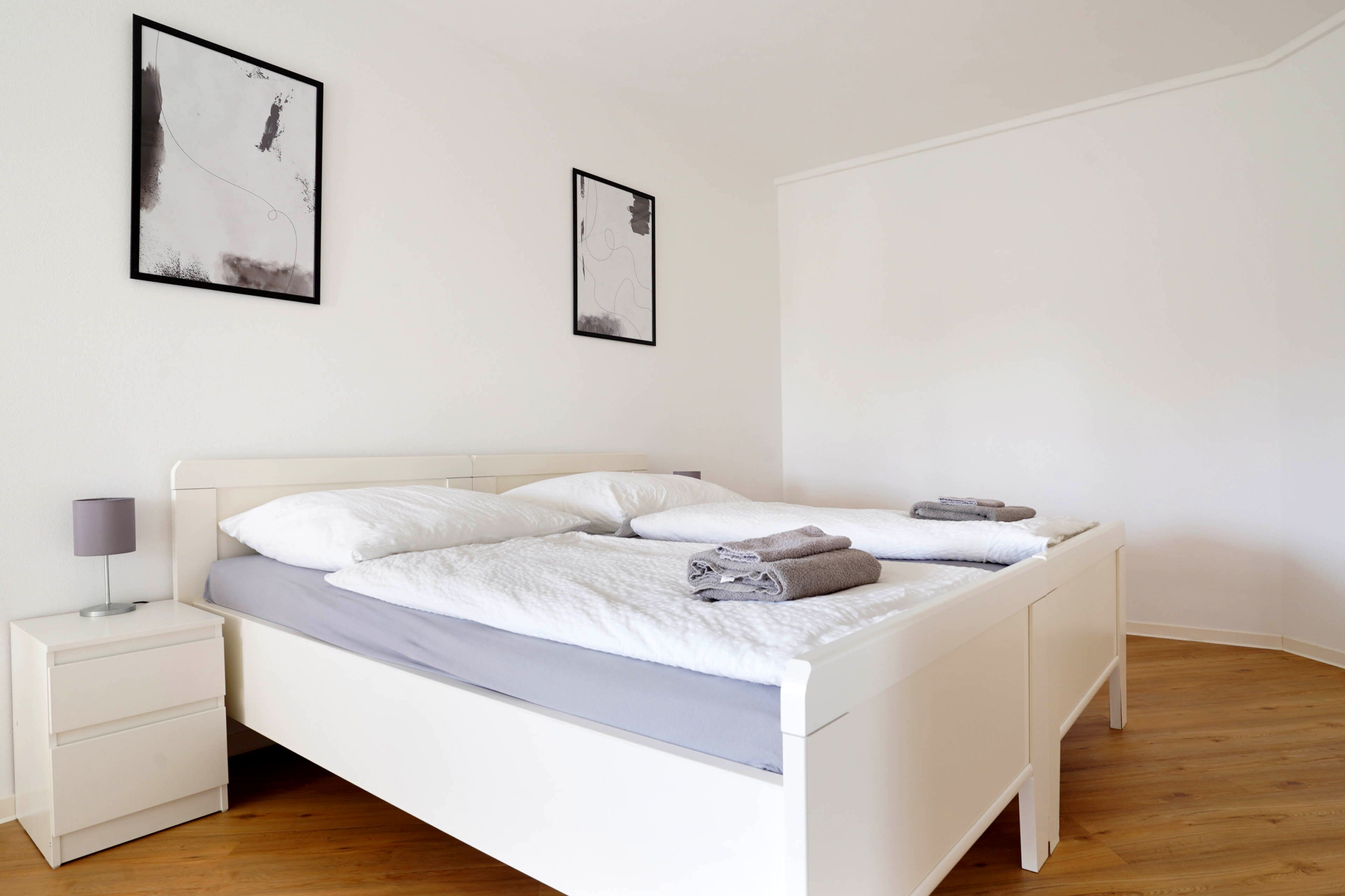 Una cama bien tendida con almohadas y toallas dobladas, una mesita de noche con lámpara, y dos obras de arte abstracto en la pared.