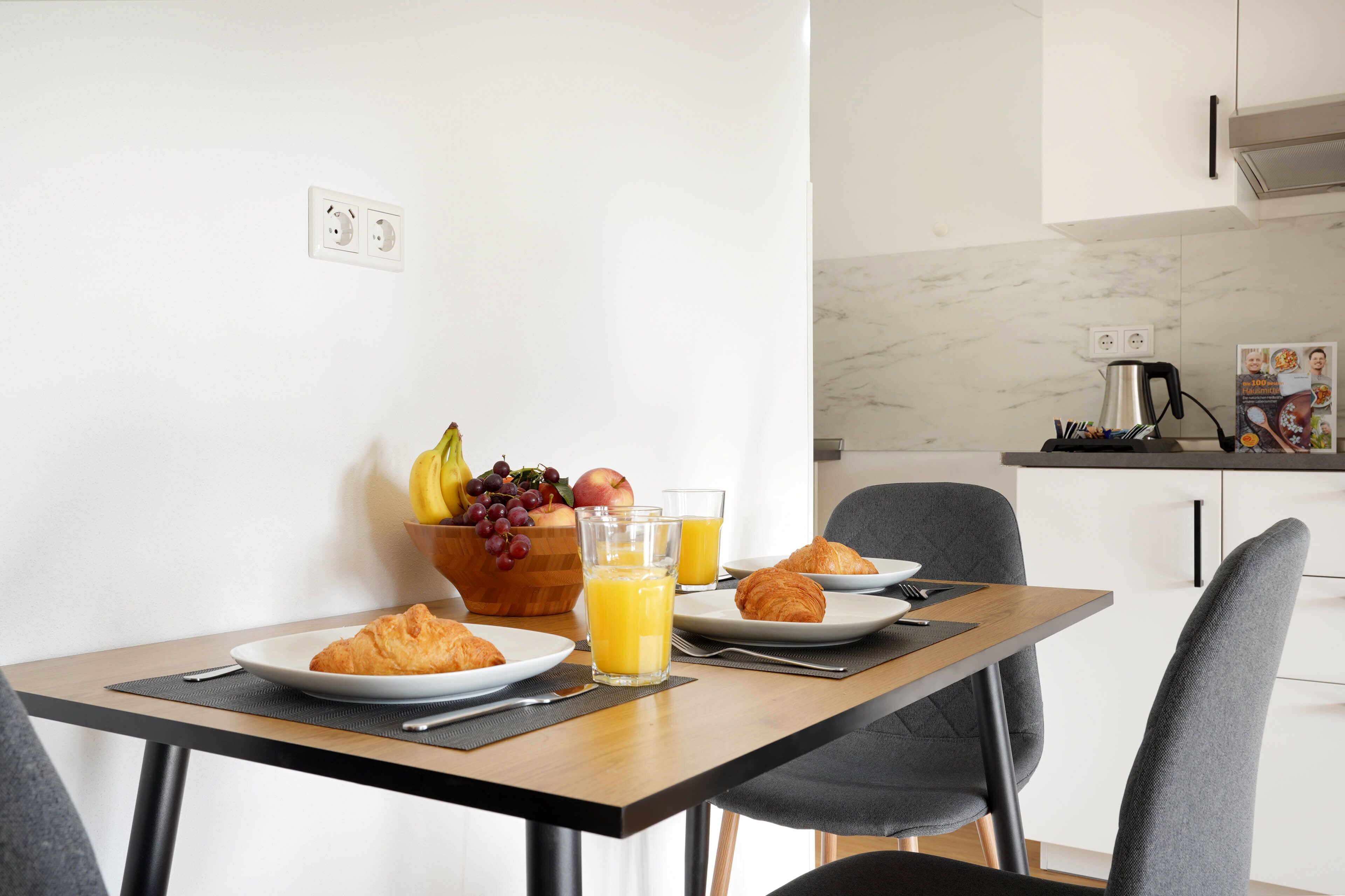 A breakfast setup with croissants, fruit, and orange juice on a table with a view of a modern kitchen in the background.