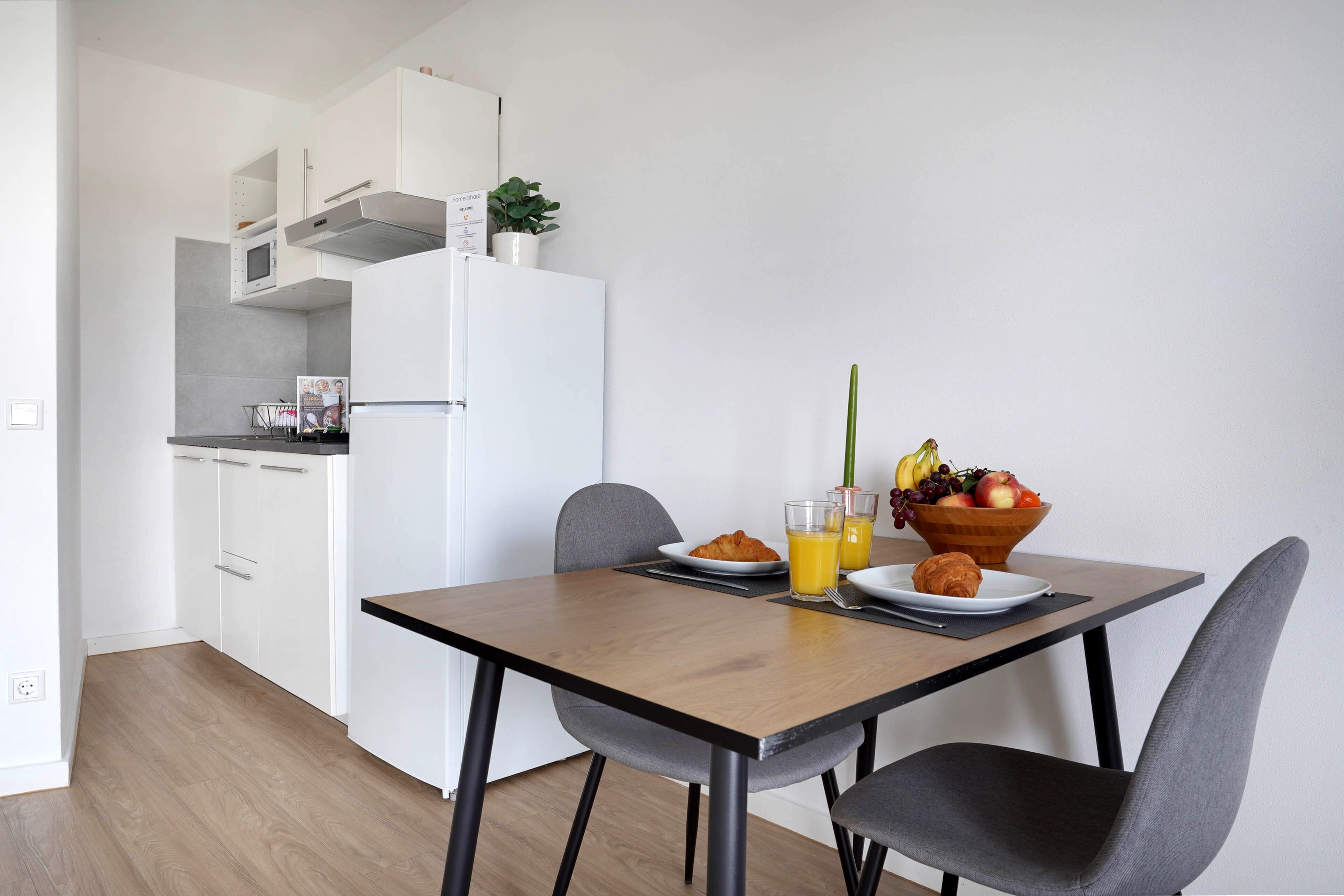 A modern kitchen with white cabinets, a microwave, and a refrigerator. There is a dining table with two chairs, a bowl of fruit, and a glass of orange juice.
