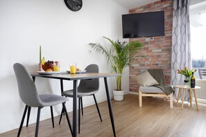 A modern living room with a dining area, a flat-screen TV mounted on a brick wall, a potted plant, and a view of the city through large windows. - Schöne Apartments in Bad Rothenfelde (Bad Rothenfelde)