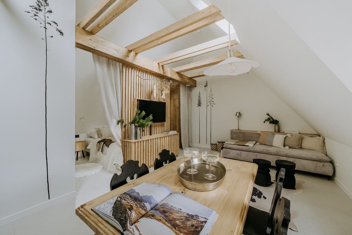 Un dormitorio estilo loft acogedor, con techo inclinado, una mesa de comedor de madera con una revista, un sofá y un televisor montado en una pared de paneles de madera.