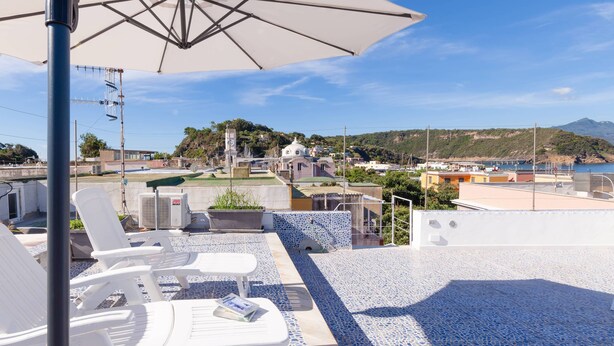 A rooftop terrace with white lounge chairs, a white umbrella, and a view of a coastal town and hills. - Da Procida 97 - Two Bedroom Apartment, Sleeps 5 (Procida)