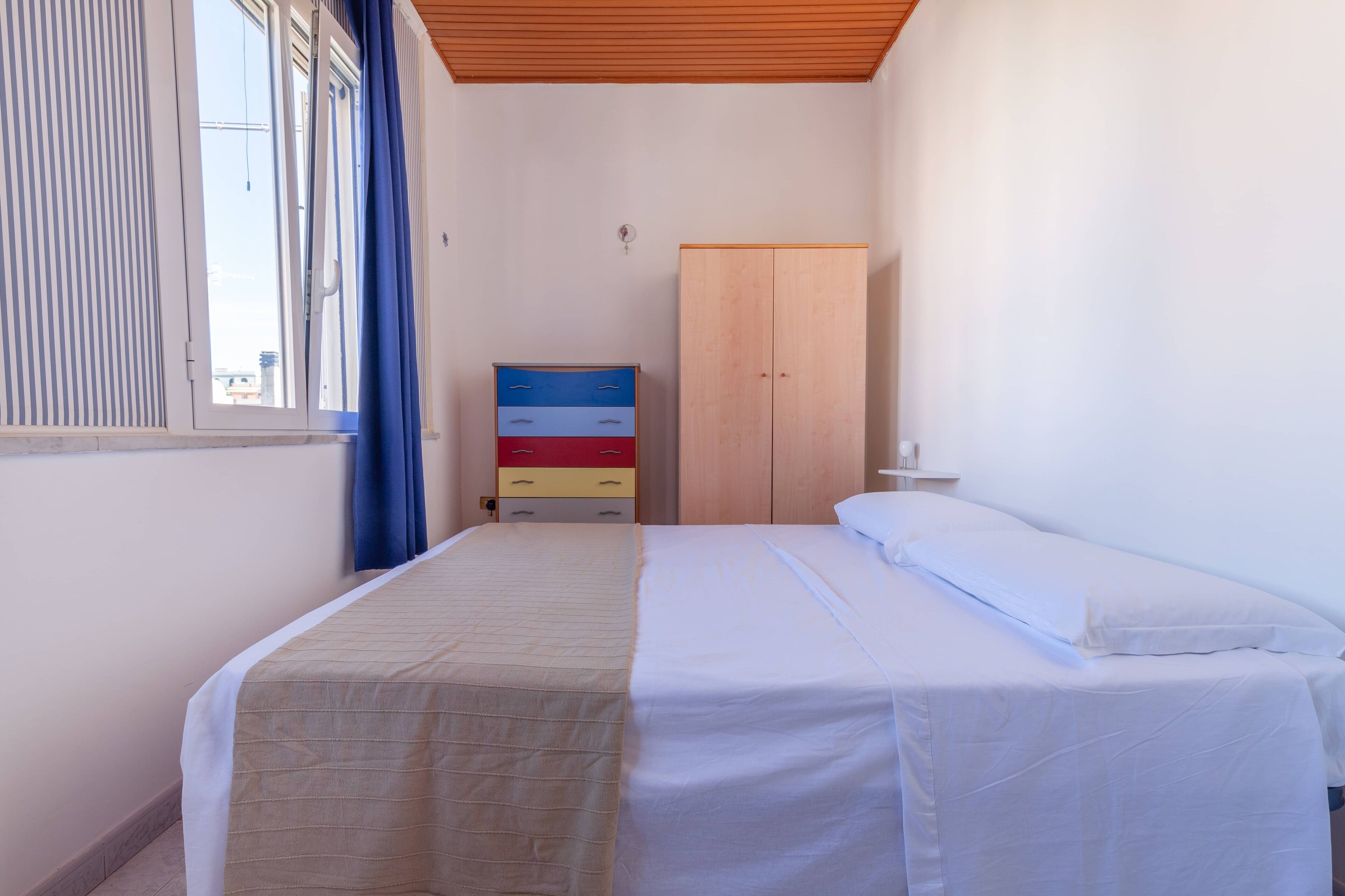 A single-bed room with a wooden ceiling, a window with blinds, a wardrobe, and a bedside table.