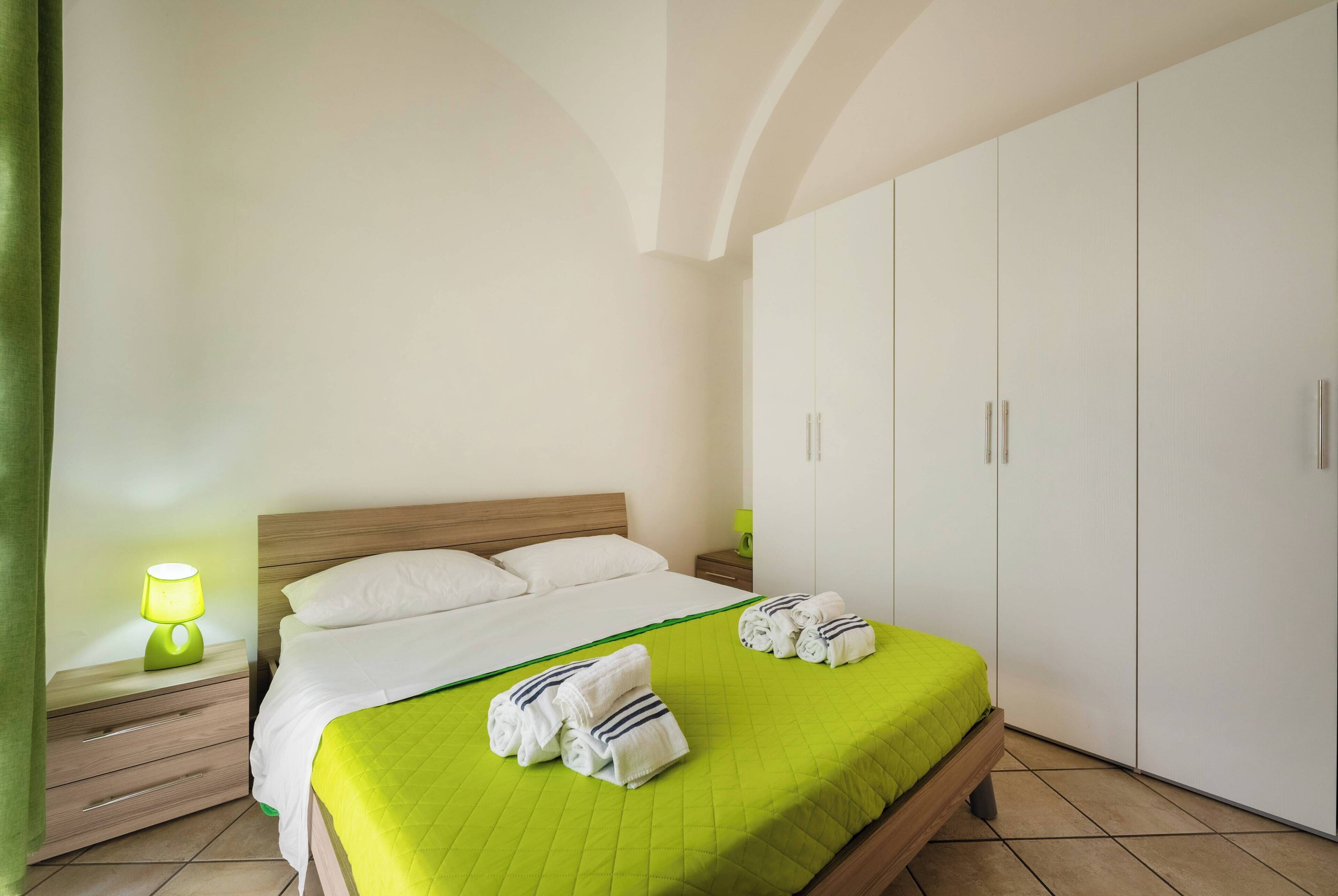 A neatly made bed with a green bedspread, a bedside lamp, and a set of towels on the floor.