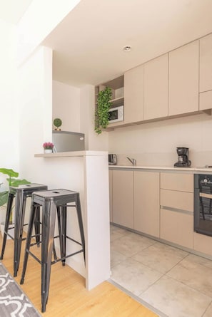 A modern kitchen with light-colored cabinets, a microwave, a coffee maker, and a dishwasher. There are two black stools and a potted plant on the counter.