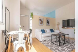 A living room with a white sofa, a black coffee table, a dining table with chairs, and a TV mounted on the wall.