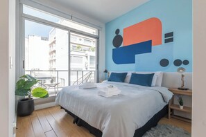 A modern hotel room with a large bed, a balcony with a view of buildings, and a colorful abstract wall art.