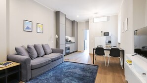 A modern living room with a grey sofa, a dining area with a table and chairs, and a kitchen in the background.