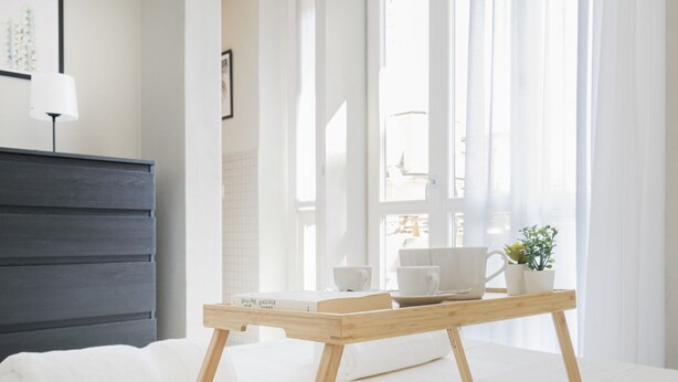 A bright room with a bed, a bedside table with a tray of items, a dark-colored dresser, and sheer curtains.