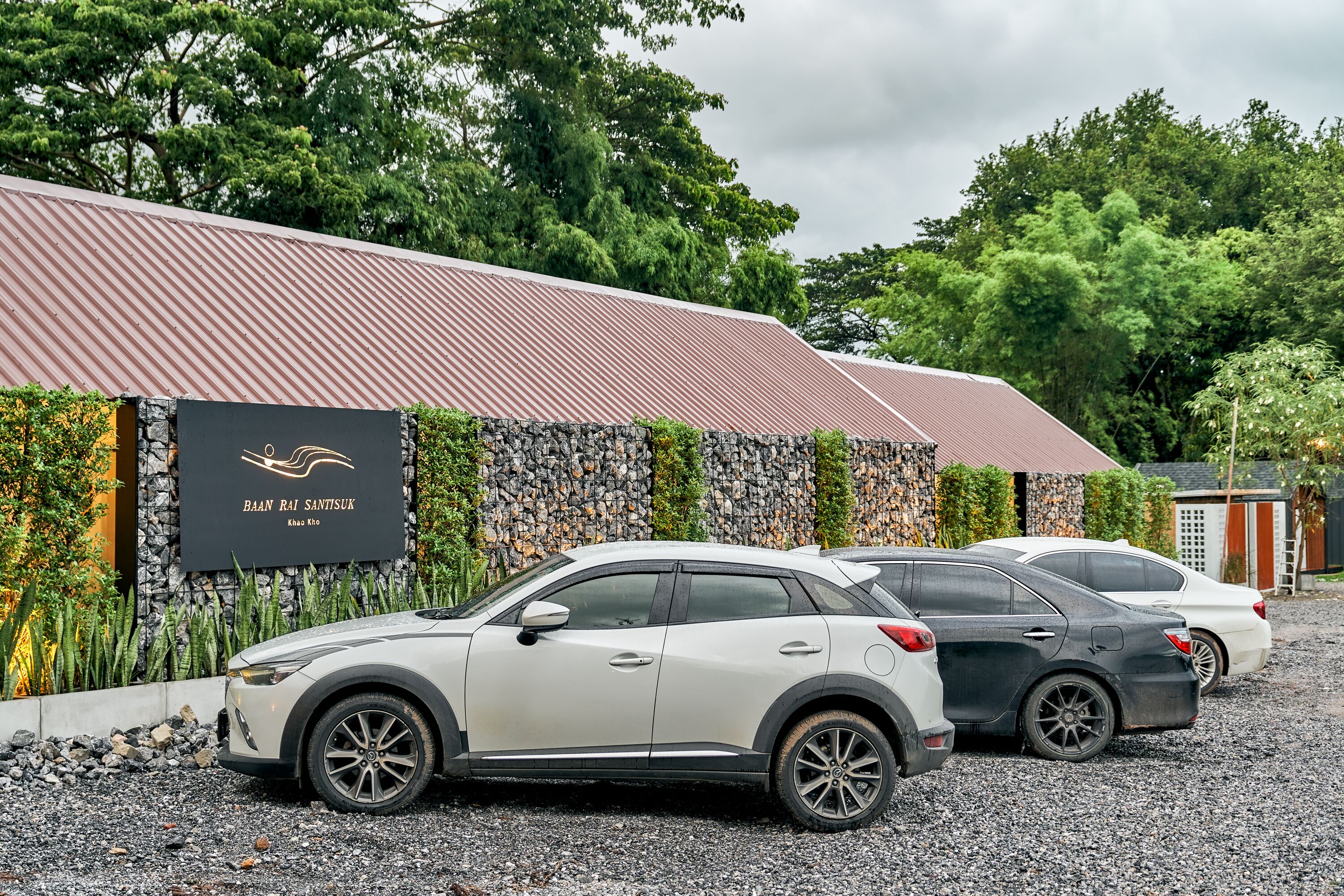 A parking area with three cars parked in front of a building with a stone wall and a sign that reads "BAAN RAI SANTISUK."