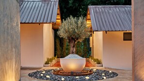A courtyard with a central water feature, surrounded by a circular stone path, and flanked by two buildings with sloped roofs. - Baan Rai Santisuk (Lom Sak)