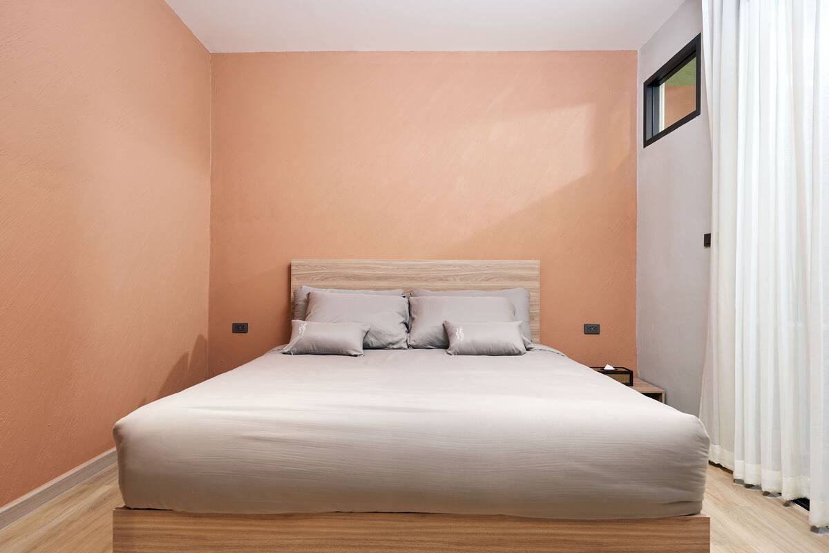 Un dormitorio con cama, almohadas, una mesita de noche y una ventana con cortinas.